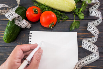 Female hand writes in a blank notebook. assorted vegetables, measuring tape and notepad on a wooden...