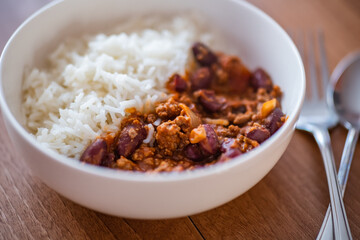 Close up and selective focus of meat chilli concarne and white rice in a white bowl on a wooden surface with intentional shallow depth of field and bokeh
