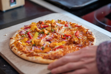 Close up and selective focus of a home cooked thin crust sweet chilli chicken pizza on a wooden cutting board being sliced with a pizza roller by an unidentifiable woman