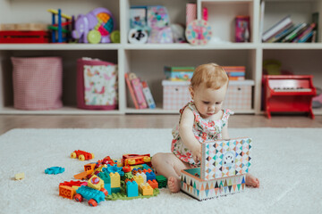 Fototapeta na wymiar Little curious baby girl playing on a playground in a bright sunny room. Learning and playing colorful educational blocks game and puzzle