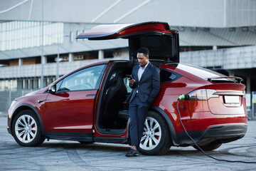 Full length portrait of successful businessman in suit using modern smartphone for checking working emails while leaning on red luxury car. African man waiting while his electric car charging.