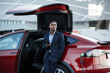Confident african man in business suit standing near luxury car with modern smartphone in hands. Handsome male using cell phone while charging battery of his red electric vehicle. - obrazy, fototapety, plakaty