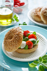 Bagel with caprese salad; cherry tomatoes, mini mozzarella cheese and basil leaves drizzled with olive oil served on a white plate close up view.