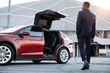 Successful african businessman holding suitcase while standing near electric car with charging...