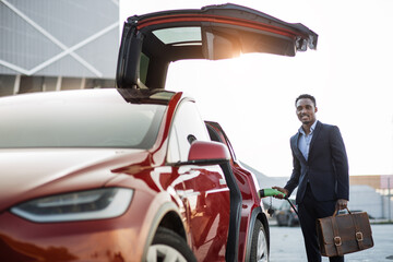 Full length portrait of handsome african man in business suit holding suitcase and charging electric car outdoors. Concept of people, modern lifestyles and eco friendly vehicle.