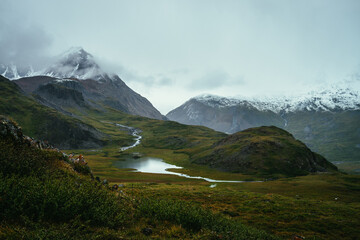 Atmospheric alpine landscape with beautiful mountain lake among rocks and great mountains with snow in overcast weather. Low cloud in highland. Rainy weather in high mountains. Snowbound mountains.