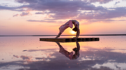 graceful gymnast performs a flexible pose at sunset in the reflection of the water beautiful...