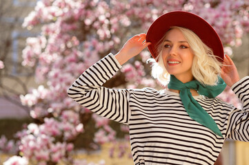 Elegant happy smiling woman wearing burgundy hat, striped longsleeve shirt, green neck scarf, posing near blooming magnolia tree. Spring fashion, lifestyle conception. Copy, empty space for text