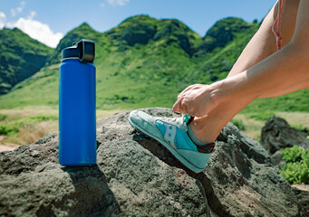 Female hiker tying her shoe next to bottle of water getting ready for and an adventure hike up a mountain. Active lifestyle, and drinking water health concept.  - obrazy, fototapety, plakaty