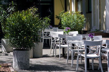 outdoor cafe terrace with white wooden chairs and tables with glass vase of flowers and concrete...