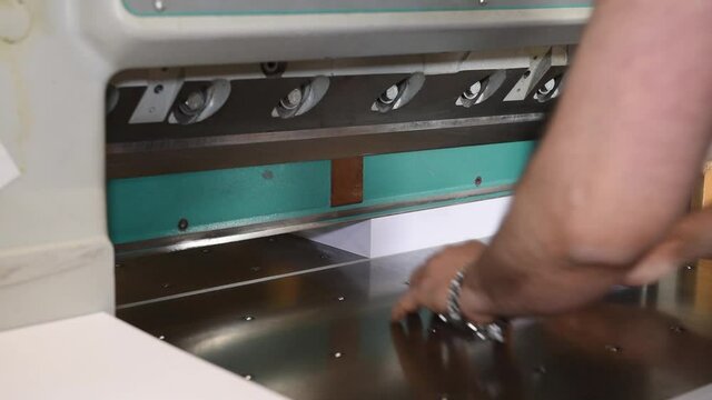 A production worker cuts a stack of printing paper on an industrial printing cutting machine in the production room. Working with your hands at the machine