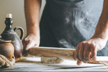 Male hands roll dough and flour with rolling pin next to clay pot and oil bottle on dark table, while cooking