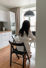 Young woman in comfortable white robe works on laptop while sitting at table the kitchen. Work at home for breakfast.