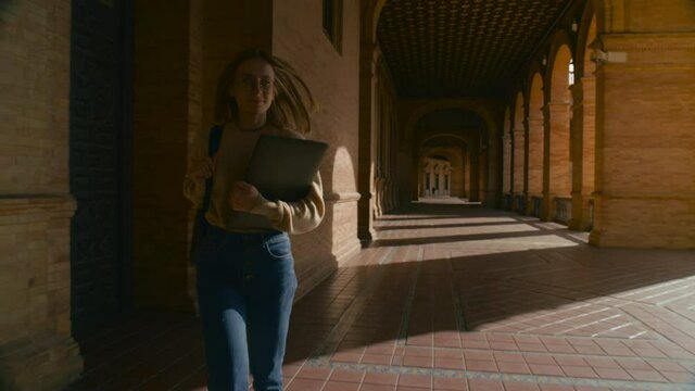 Excited young female student walk for first time through corridors of red brick university campus. Beautiful happy woman, ready to learn and gain new experience. Higher education abroad or exchange