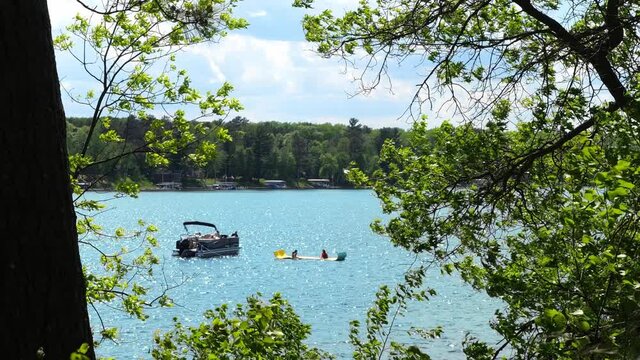 Children, on a floating device tied to a pontoon on a beautiful lake, are seen enjoying their day, through tree branches on a sunny day. Boating in Minnesota. Subtle zoom out.