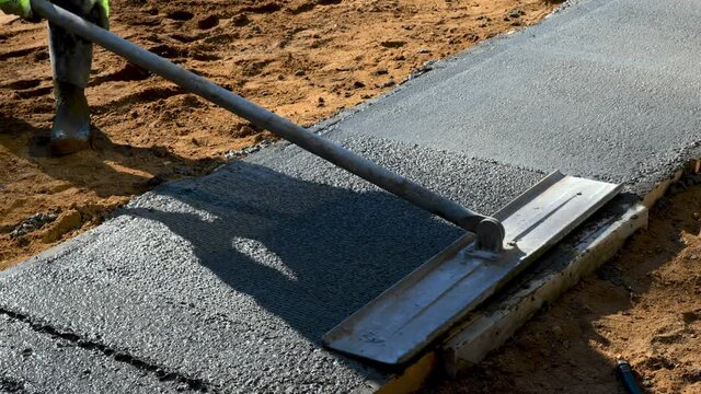 Closeup clip of a bull float being used on new cement concrete by a worker to bring liquid to the surface and to level, and smooth the new sidewalk at a construction job site.