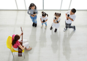 Top view shot of Asian female teacher playing guitar teaching group of little young kindergarten boy and girls student holding microphone in hands learning to sing a song in music classroom studio