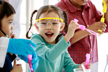 Portrait closeup shot of Asian little playful happy pre-elementary schoolgirl with bunches braided pigtail hairstyle wears safety goggles playing blowing soap bubbles maker with friends in laboratory