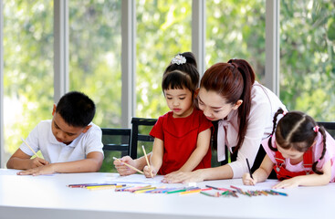 Fototapeta premium Asian female pretty single mother standing helping teaching little cute innocent daughter drawing and painting with colored pencils on paper book while other boy and girl doing homework together