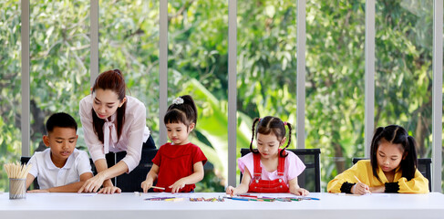 Asian female primary school tutor teacher teaching smart clever boy student do homework while other little girls writing drawing painting on paper book with colored pencils together on table at home