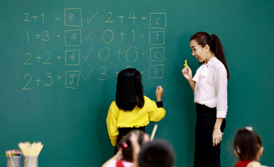 Portrait shot of Asian beautiful female mathematic teacher standing smiling cheer up while little smart schoolgirl student solving math equations on chalkboard in front of classmates in classroom