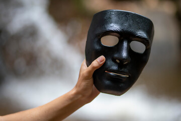 Select the focus of the black mask on the Businessman's hand holding it. Nature waterfall background. Concept. Take off the mask, reveal your identity, and return to nature. Take off the dark side