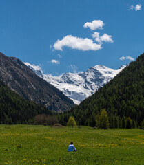 Fototapeta na wymiar Lonely man sitting in the middle of a green mountain meadow, full of yellow flowers, admiring the beauty of a snowy mountain range overhung by blue sky