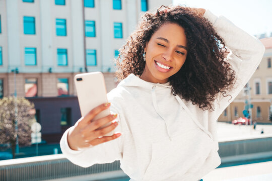 Beautiful black woman with afro curls hairstyle.Smiling hipster model in yellow hoodie. Sexy carefree female posing on the street background in sunglasses. Cheerful and happy.Taking selfie photos