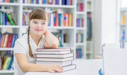 Pensive young girl with syndrome down sita at library with books. Empty space for text. Education for disabled children concept