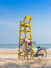 Fototapeta premium Old retro black bicycle with flowers bouquet beside Life ring for life safety on yellow lifeguard stand station or lifeguard chair protecting the safety of tourist on the empty beach in sunshine.
