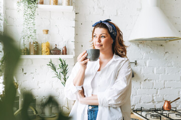 Attractive young woman plus size body positive in blue jeans and white shirt drink morning coffee at the home kitchen