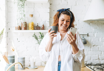 Attractive young woman plus size body positive in blue jeans and white shirt using cellphone at the home kitchen - Powered by Adobe