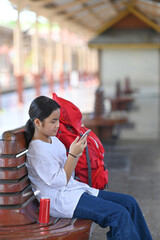 Asian girl with backpack sitting in railway station and using smart phone. Holiday, journey, trip and travel concept.