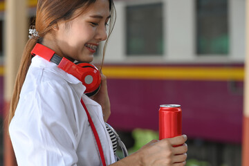 Smiling young female wearing headphone and holding refresh drink wile standing in railway station.
