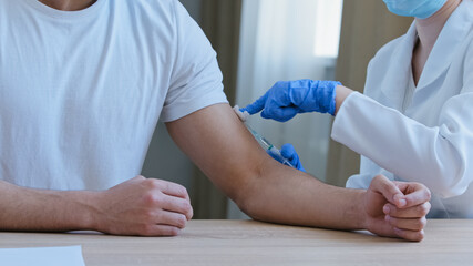 Close-up unrecognizable doctor in protective medical uniform makes two injections with syringe and needle to man in shoulder changes medicine does vaccination immunization sitting at table in clinic