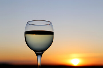 Glass with white wine on beautiful sunset background, sun and sky are reflected in alcohol drink. Concept of celebration, evening party at resort, romantic dinner outdoors