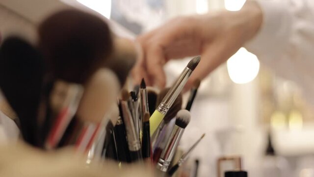 Crop makeup artist choosing brush. Unrecognizable female professional visagiste picking applicator from collection placed on table in beauty studio 