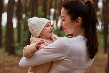 Beautiful dark haired female wearing white clothing posing outdoor, holding infant baby in hands and looking at daughter with great love, playing in forest, smiling happily.