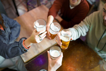Gathering of best friends sitting in the pub rising beer glasses clinking in a celebratory toast. Young people having fun drinking alcoholic drinks in the night concept