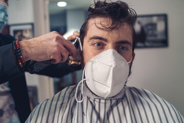 Barber cutting hair with a clipper to a young guy with a ffp2 face mask during the coronavirus...