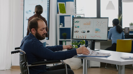 Disabled immobilized invalid businessman working at financial project respecting deadline typing on computer analysing statistic reports from documents sitting in wheelchair in start up office