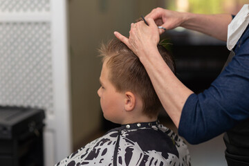 Cutting hair for young boy in a beauty salon.Hairdresser's hands holding a comb and a colorful scissors.Closeup.