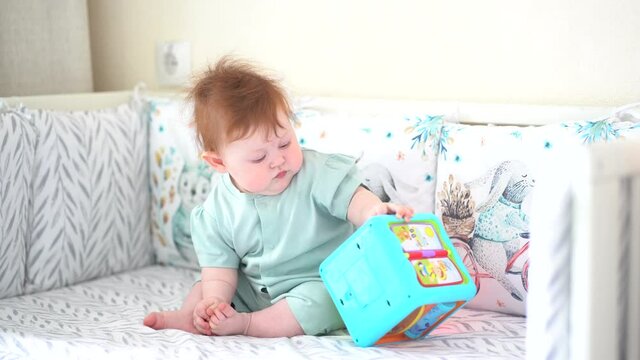A baby with red hair sits in a crib and plays with a musical toy
