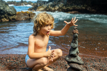Funny kid boy making stack of pebbles stone against sea background for spa, balance, meditation and zen theme. Child play on the beach.