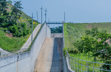 Dry concrete spillway on rural hillside covered with grass and trees.