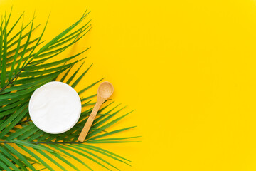 Cosmetic cream with a wooden spoon and a palm branch on a yellow background. Spa care concept. Horizontal orientation, copy space.