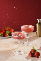 Two glasses with red strawberry dessert with ice cubes, crashed ice and water splashes on burgundy background
