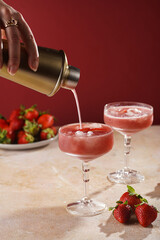 Female hand pouring cocktail our of shaker into a glass with red strawberry dessert with ice cubes and crashed ice and strawberry slices on burgundy background