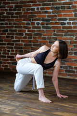 Fototapeta na wymiar Beautiful young woman with white sporty pants and black top practicing hatha yoga Parivrtta Anjaneyasana pose crescent lunge on knee with twist, against the background of a brick wall in the loft. 