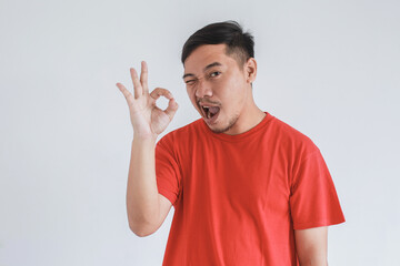 Fototapeta premium Funny face expression of Asian man wearing red t-shirt with wink and OK gesture in his hand